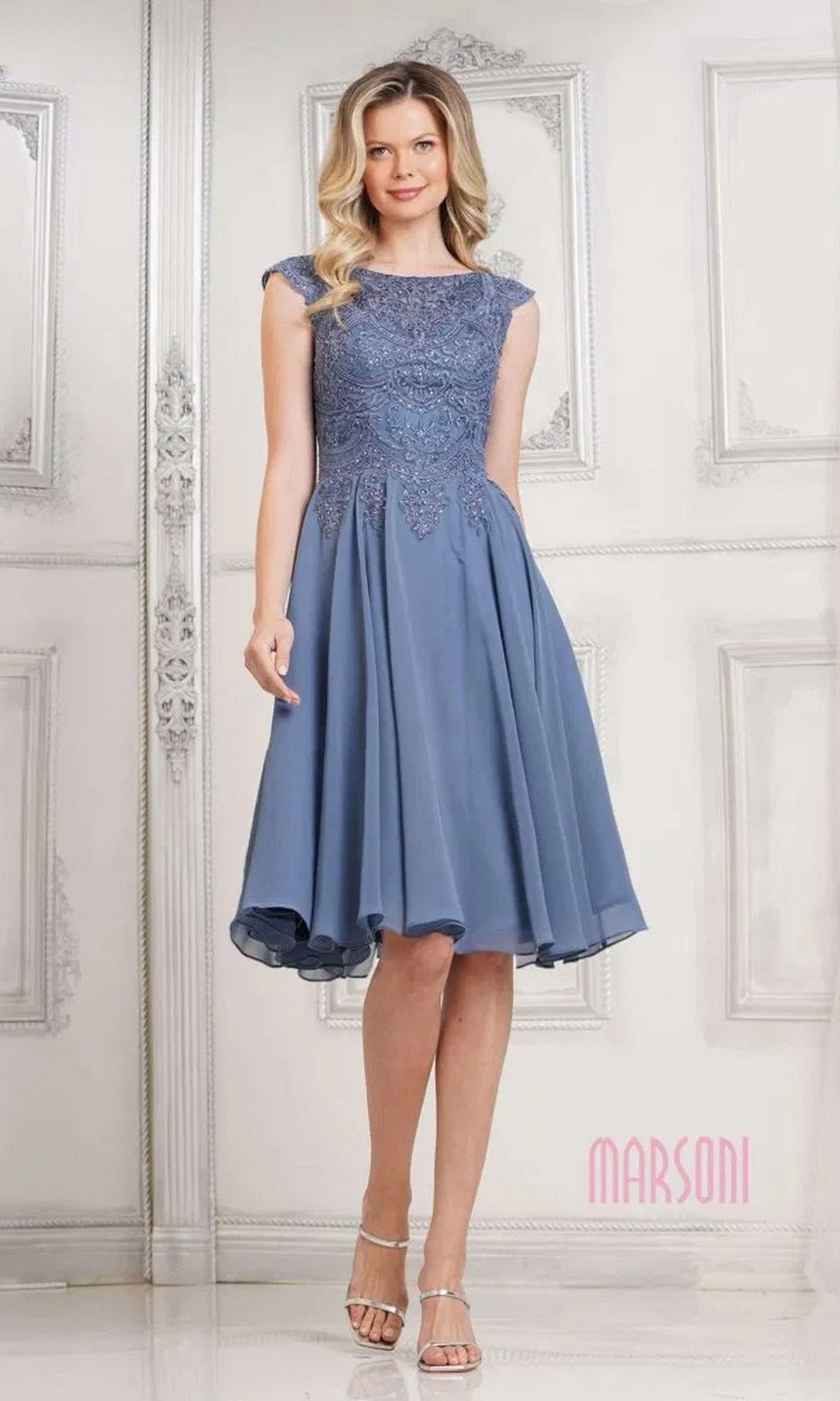 A woman in a Marsoni M238S knee-length semi-formal dress in slate blue with a lace bodice and chiffon skirt, paired with silver heels, stands before a white decorative wall.